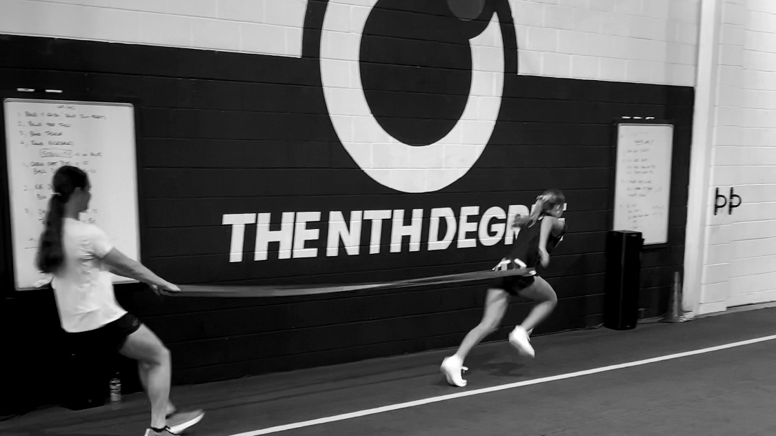 Two athletes mid resisted-sprint inside the Nth Degree training room, motion blur on the runner.