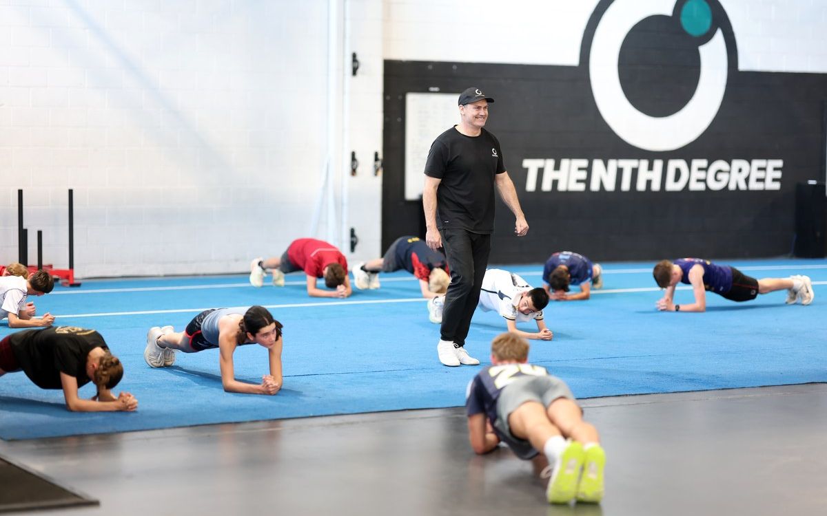 A group of athletes holding a plank on the blue turf at The Nth Degree, Norwood, with a coach walking among them.