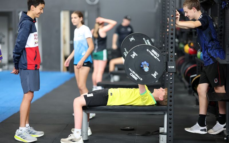 An athlete bench pressing a loaded barbell in the rack while teammates look on.