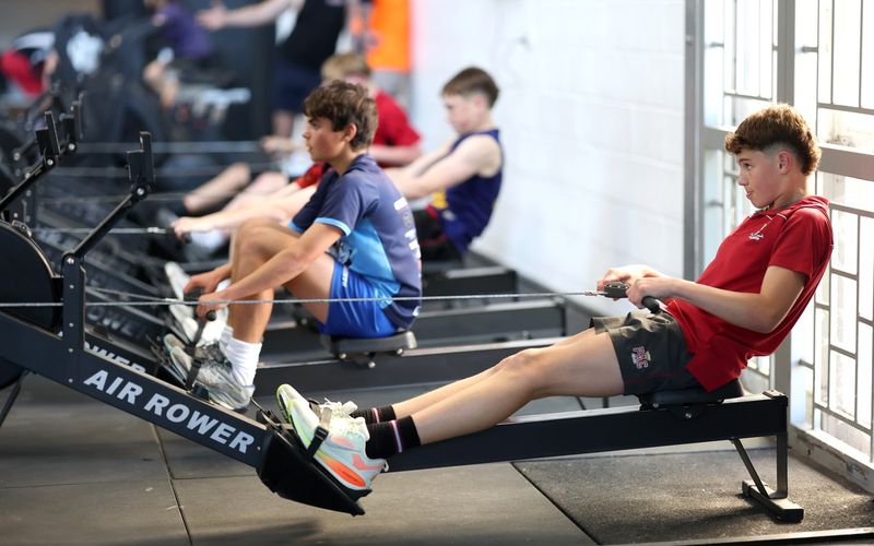 A row of athletes working on the air rowers by the windows.